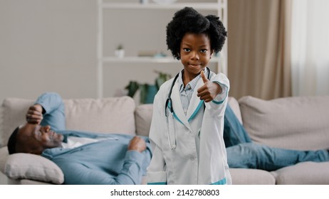 Little African American Daughter Playing With Dad Father Patient Lying On Couch Kid Girl Pretending To Be Doctor Looking At Camera Smiling Showing Thumb Up No Problem Excellent Health Approval Gesture