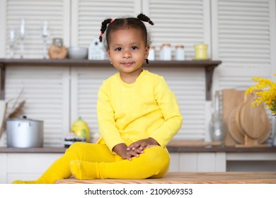 A Little African American Girl In A Yellow Dress With Curly Pigtails Sits In The Kitchen.