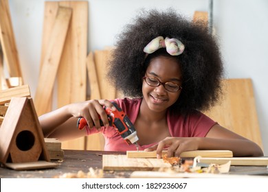 A Little African American Inventor Practice To Be A Carpenter In Carpentry Shop. A Girl Working As Woodworker To Drill Holes In A Wooden Plank At Workshop. DIY Woodworking Crafts And Hobbies Concepts