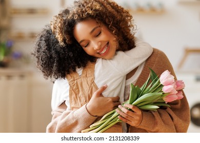 Little African American Son Congratulating Mom With Mother's Day At Home And Giving Her Fresh Flower Bouquet, Happy Mixed Race Woman Mother Embracing Son. Family Holidays And Celebration Concept