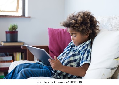 Little African Boy Sitting On Sofa And Playing Game On Digital Tablet. Portrait Of A Young Black Child At Home Watching Cartoon On The Laptop. Modern Kid And Education Technology.