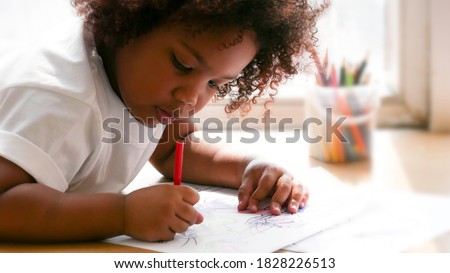Little African girl painting and drawing on paper. Preschool girl lying writing on floor.
