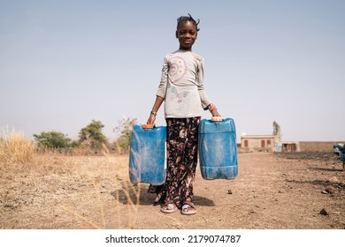 Little African Girl Standing Not So Far From Her Poor Parents' Home With Two Water Canisters In Her Hands, Symbolizing The Hard Work Of Household Water Supply