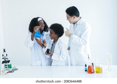 Little African Kids Studying Chemistry And Doing Chemical Science Experiment In Laboratory At School. Happy Dark Skinned Girl Showing Test Bottle To Asian Teacher Man And He Thumbs Up