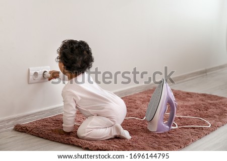 Little African-American baby playing with socket and iron at home. Child in danger