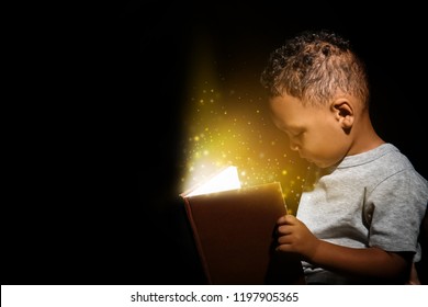 Little African-American boy reading magic book on dark background