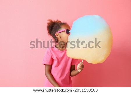 Little African-American girl eating cotton candy on color background