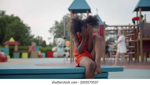 Little Afro Girl Sitting Alone On Bench In Summer Park And Crying. Portrait Of Unhappy Preschool Afro-american Child Sitting Alone On Playground