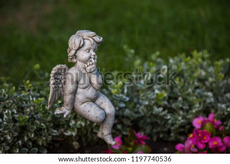 A little angel has his forefinger in his mouth and sits on a bush