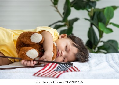 Little Asian Baby Girl Lying Down Sleeping On Bed At Home, Hugging Teddy Bear Doll, With American Flag. Newborn Child Relaxing In Bed. Nursery For Young Children.