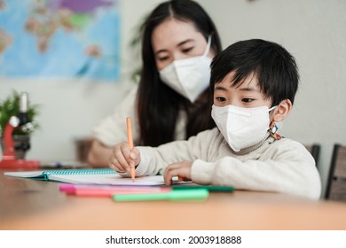 Little Asian Boy Drawing With Nanny In Background While Wearing Safety Face Mask For Coronavirus Outbreak - Daycare And Pandemic Concept