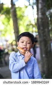 A Little Asian Boy Eating Sausage With A Bamboo Stick. 