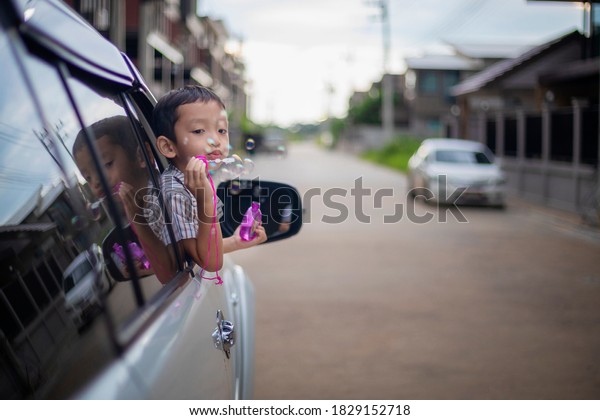 A little Asian boy happily blows bubbles out of a\
car window.