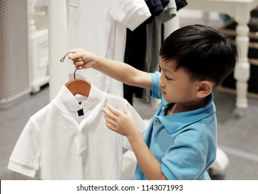 The Little Asian Boy Holding Coat Hanger And Checking Polo Shirt. Background With Many Kid Shirt Hanging In Closet. Concept Of Buy, Sell And Kid Clothes Fashion .