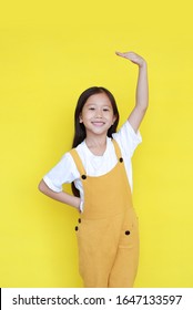 Little Asian Child Girl Measures The Growth On Yellow Background. Kid Estimate Her Height By Hand With Looking Camera