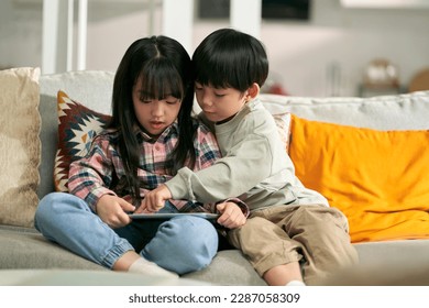 little asian children brother and sister sitting on family couch at home using digital tablet computer together - Powered by Shutterstock