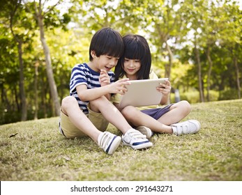 Little Asian Girl And Boy Sitting On Grass Using Digital Tablet Outdoors In A Park.