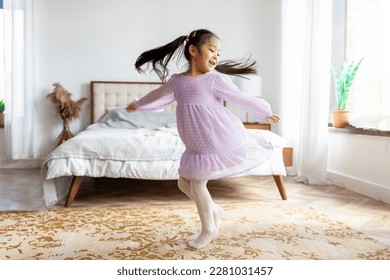 little Asian girl in dress dances and spins at home, Korean child in festive outfit moves and rejoices in the room near the bed - Powered by Shutterstock