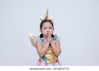 A little asian girl dressed in a colorful unicorn costume with a golden horn and wings, pouting and saying please, with hands clasped begging. Isolated on a white background. - Powered by Shutterstock