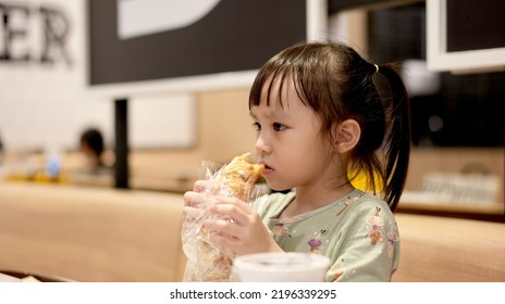 Little Asian Girl Eating Bread At Restaurant., Background. Copy Space.