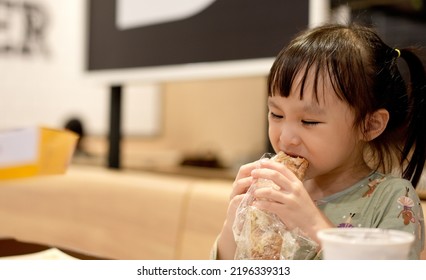 Little Asian Girl Eating Bread At Restaurant., Background. Copy Space.