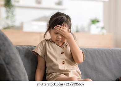 Little Asian Girl Have A Head Ache On Couch At Home. Child Touching On Head Have A Fever Higher Temperatures So Pain And Illness. Kid Has A Grimace Face. Child Health Care