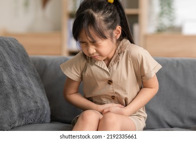 little Asian girl have a stomach ache on couch at home. Child squeeze on her belly so pain and illness. kid has a grimace face. Child health care