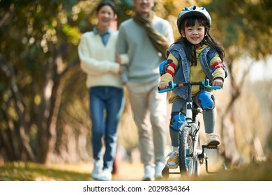 little asian girl with helmet and full protection gears riding bike in city park with parents watching from behind