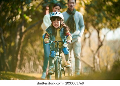 little asian girl with helmet and full protection gears riding bike in city park with parents watching from behind