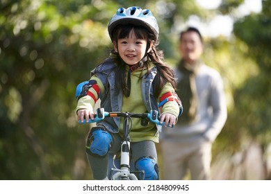 little asian girl with helmet and protection gear riding bike in city park with father watching from behind - Powered by Shutterstock