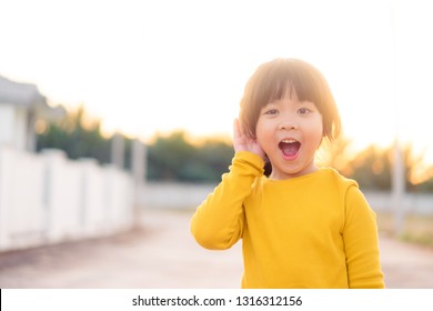 Little Asian Girl Holds Her Hand Near Her Ear And Listening Something.Exciting Face On Funny Child Girl Wear Yellow Shirt In Winter Time And Listening To Curious Good News. 