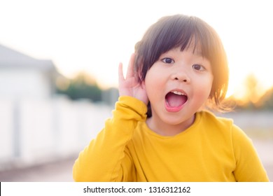 Little Asian Girl Holds Her Hand Near Her Ear And Listening.Disability Day.Deaf Kid.Exciting Face On Asian Child Girl Wear Yellow Shirt In Winter Time.Turning Off Phone And Listening Your Child.