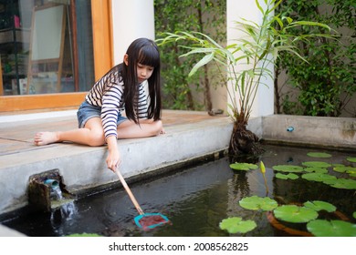 Little Asian Girl With Long-haired And Bangs Sitting On Floor Beside The Pond Using A Scoop Net Catching A Small Fish In The Water Pond With Lotus Leaves, Running Water And Plants In The Home Backyard