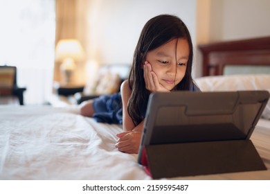 Little Asian Girl Is Lying Down On The Bed In The Bedroom And Looking The Screen Of Tablet Alone With Happy Moment, Concept Of Internet, Social Media, Online And Technology Impact To Children Life.