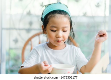 Little Asian Girl Make Boring Face When She Eat Breakfast.Unhappy, Disgusted, Unlike Food In The Morning Concept.Little Asian Girl Child With No Appetite In Front Of The Meal.