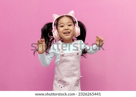 little asian girl in pink children's headphones listens to music and dances on pink isolated background, korean child in pink sundress and unicorn headphones jumps and rejoices
