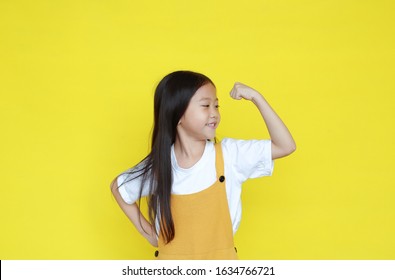 Little asian girl raising hand up showing his muscle isolated on yellow background. Child in dungarees with good health concept