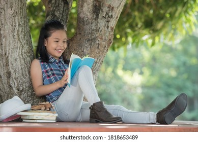 Little Asian Girl Reading A Book Under Big Tree With Hat, Book, And Model Plane Beside. Children And Science. Blurred Background. Learning The Imagination And Dreams Of Rural Child. Studying At Home.