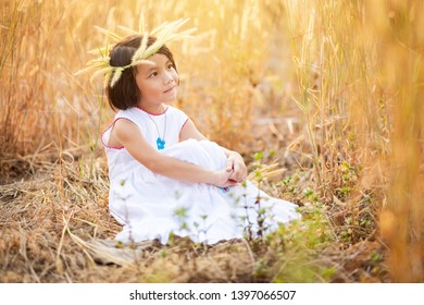 Little Asian girl sitting on ground in grass field, Kid in white dress and flower crown sitting alone in the field with warm sunlight in the evening - Powered by Shutterstock