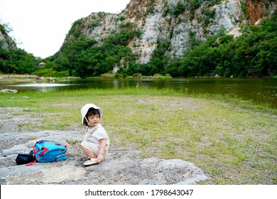 A Little Asian Girl Traveler Sighseeing And Sit On The Rocks.Toddler Sit Next To Her Small Bagpack On A Beautiful Lake Background Of Mountains Called Kao Ngu Rocks Park. Travel With Kids. Family Trips