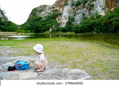 A Little Asian Girl Traveler Sighseeing And Sit On The Rocks.Toddler Sit Next To Her Small Bagpack On A Beautiful Lake Background Of Mountains Called Kao Ngu Rocks Park. Travel With Kids. Family Trips