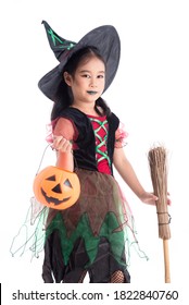 Little Asian Girl In Witch Costume With Make Up Holding Pumpkin Bucket And Broom Standing Over White Background. Child Halloween Costume Concept.