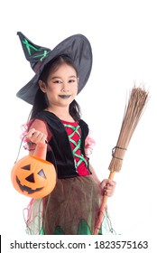 Little Asian Girl In Witch Costume With Make Up Holding Pumpkin Bucket And Broom Standing Over White Background. Child Halloween Costume Concept.