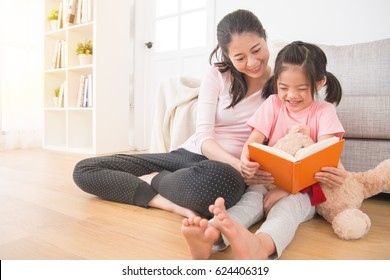 little asian happy girl holding teddy bear watching new comic book with the beautiful mixed race mom sitting together on floor enjoy the holiday afternoon time, family home activity concept.