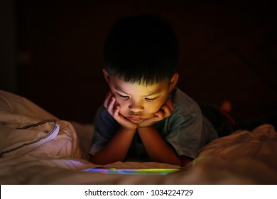 Little Asian Kid Alone Watching Tablet Device, Lying On White Duvet Bed With Chin On Hands, In Background Darkness Bedroom Night Time. Colourful Bright Light From Screen Reflex On The Boy Face.