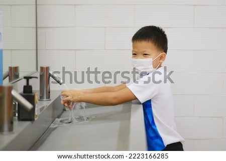 Little Asian student kid wearing medical face mask washing hands by himself on sink  in the basin, bathroom sink and water drop from faucet in public toilet, Clean school washrooms, Selective focus