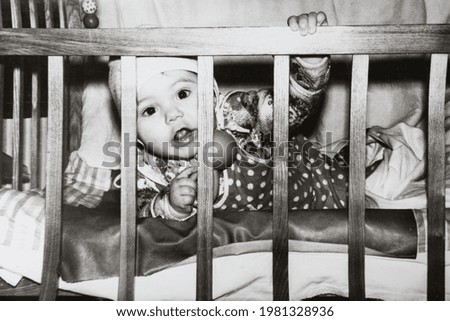 Little baby in bed looking through the wooden rods. Vintage black and white paper photo. Early 1980s. Old surface, soft focus. Transferred property, family archive.