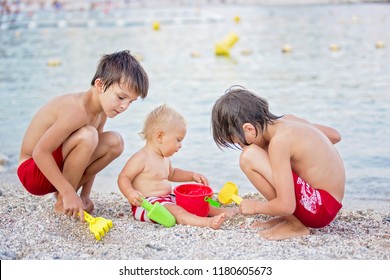 Little Baby Boy And His Older Brothers, Playing On The Beach With Plastic Toys And Pebbles