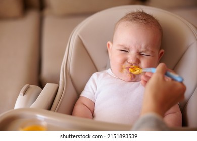 Little Baby Boy Sitting In High Chair Making Funny Faces And Refusing To Eat Pumpkin Porridge While Mother Trying To Feed Him Using Spoon