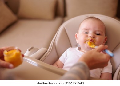 Little Baby Boy Sitting In High Chair Making Funny Faces And Refusing To Eat Pumpkin Porridge While Mother Trying To Feed Him Using Spoon